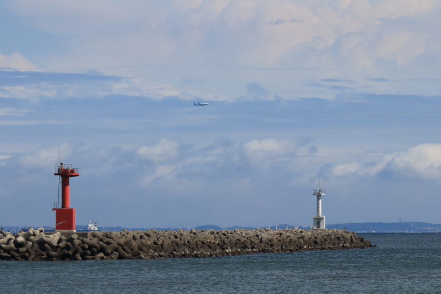 海にテトラポッドの防波堤と赤と白の2つの灯台があり空には飛行機が飛んでいる画像
