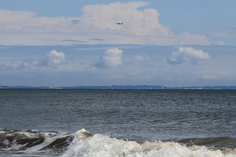 砂浜に打ち付ける波と海と白い雲が浮かぶ空には飛行機が飛んでいる画像