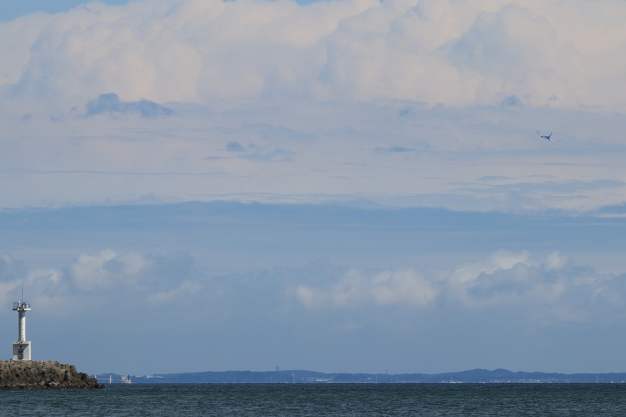 海にテトラポッドの防波堤と白の灯台があり空には飛行機が飛んでいる画像