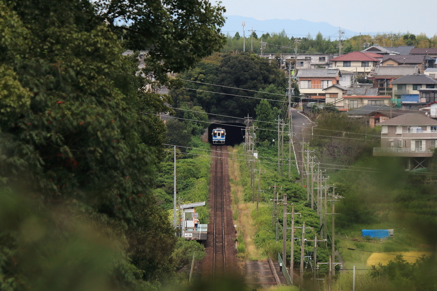 直ぐな線路と小さな無人駅には一人の人がいる。線路の先のトンネルから列車が走ってきます。線路の周囲の風景も写っているの画像