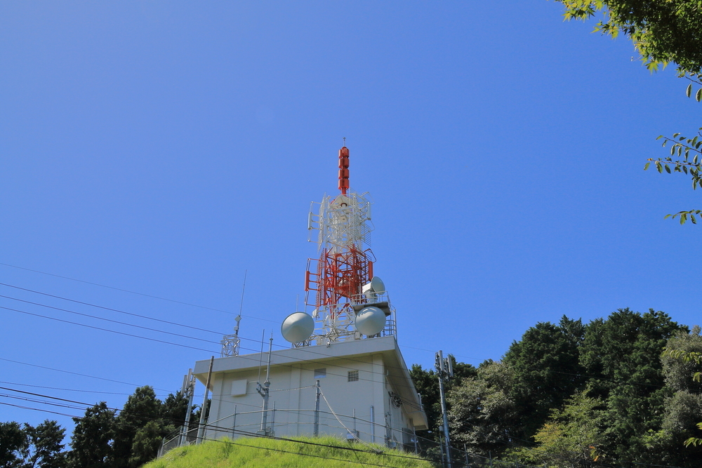 白い建物の上に白と赤の放送用の鉄塔と奥に森が映る画像