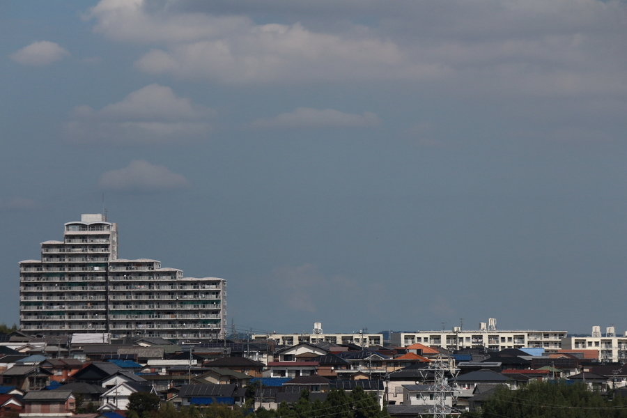 一棟の高層マンションと住宅街を高台から見下ろしています。その背後は少し霞んだ青空に雲が少し浮かんでいる画像。
